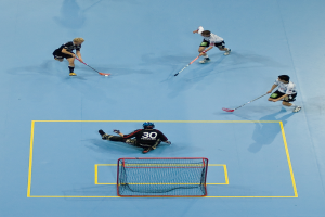 Drei Personen in Sportkleidung spielen Hockey, wobei eine Person auf der rechten Seite den Ball mit einem Hockeystock schlägt, während eine andere Person hinter einem Netz auf dem Boden sitzt.