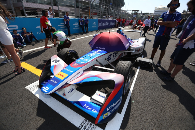 Formula E-Auto mit weißer, blauer, roter und schwarzer Lackierung auf einer Rennstrecke geparkt, umgeben von Menschen, mit einem Schirm im Vordergrund und Rennstrecken-Infrastruktur im Hintergrund unter einem klaren blauen Himmel.