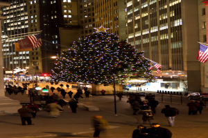 Ein mit Menschen geschm√ľckter Weihnachtsbaum in der Mitte einer Stra√Ÿe, flankiert von zwei Fahnenm√§sten, mit beleuchteten Geb√§uden im Hintergrund.