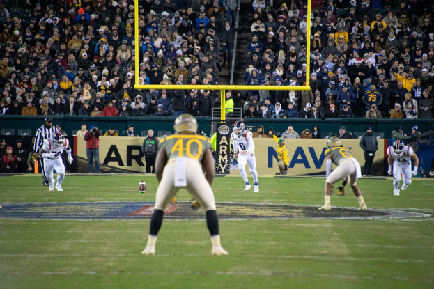 Fußballspieler in Sportkleidung und Helmen konkurrieren auf einem Feld mit Zuschauern in den Rängen und sichtbaren Stadionanzeigen.
