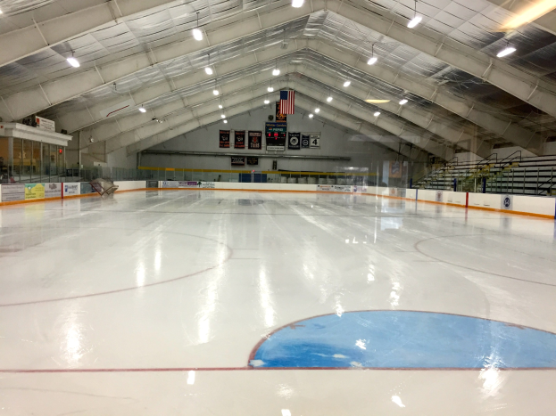 Große Indoor-Eisbahn mit Deckenbeleuchtung, Werbetafeln, einer Rückwand, Treppen mit Geländern und einigen Menschen, die einem Hockey-Sportplatz ähneln.