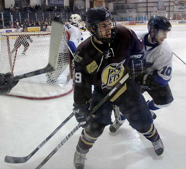 Junge Männer in Helmen und Hockey-Ausrüstung spielen Eisockey auf einer Indoor-Eisfläche mit Zuschauern in den Rängen und beleuchteten Bannern.