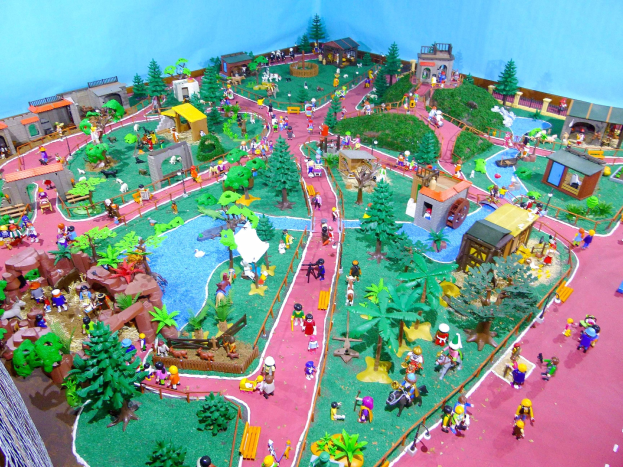 Zoo-Modell mit Miniaturfiguren, Bäumen, Häusern, Pflanzen, Gras, einem Zaun und einer Hintergrundwand, mit Text unten.