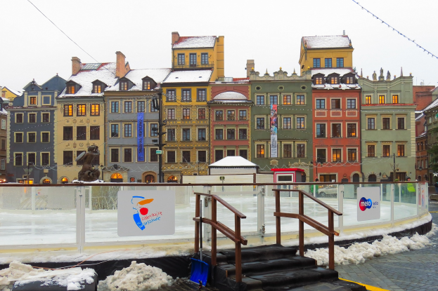 Eislaufbahn in einem Stadtplatz umgeben von Geb├Ąuden, Treppen, Gel├Ąndern, Informationsschildern, schneebedeckter Erde, Laternenmasten, B├Ąumen und Himmel.