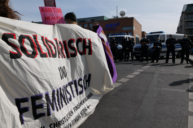 Gruppe von Menschen marschiert die Straße entlang und hält ein "Solidarität und Feminismus"-Schild, mit parkenden Fahrzeugen, Gebäuden, einer Schüsselantenne und einem klaren blauen Himmel im Hintergrund.