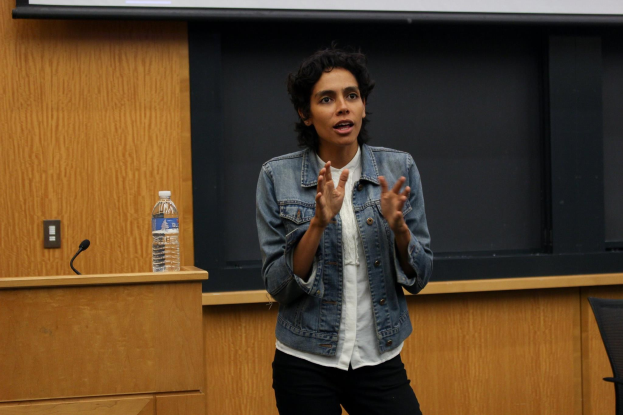 Frau am Rednerpult mit Mikrofon und Flasche, die eine Pr├Ąsentation in einem holzvert├Ąfelten Veranstaltungsort gibt, mit einem Projektionsschirm und einem Stuhl rechts von ihr.