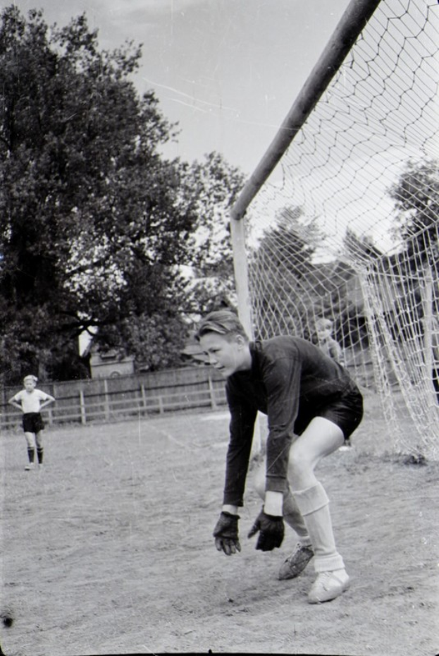 Ein Schwarz-Weiß-Foto eines jungen Jungen, der Fußball auf einem Feld spielt, mit einem Zaun, Bäumen und einem klaren Himmel im Hintergrund, anderen Kindern im Hintergrund und einer Personenschuhwerk im Vordergrund.