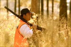 Eine Person mit einer Waffe steht vor Pflanzen mit Bäumen im Hintergrund.