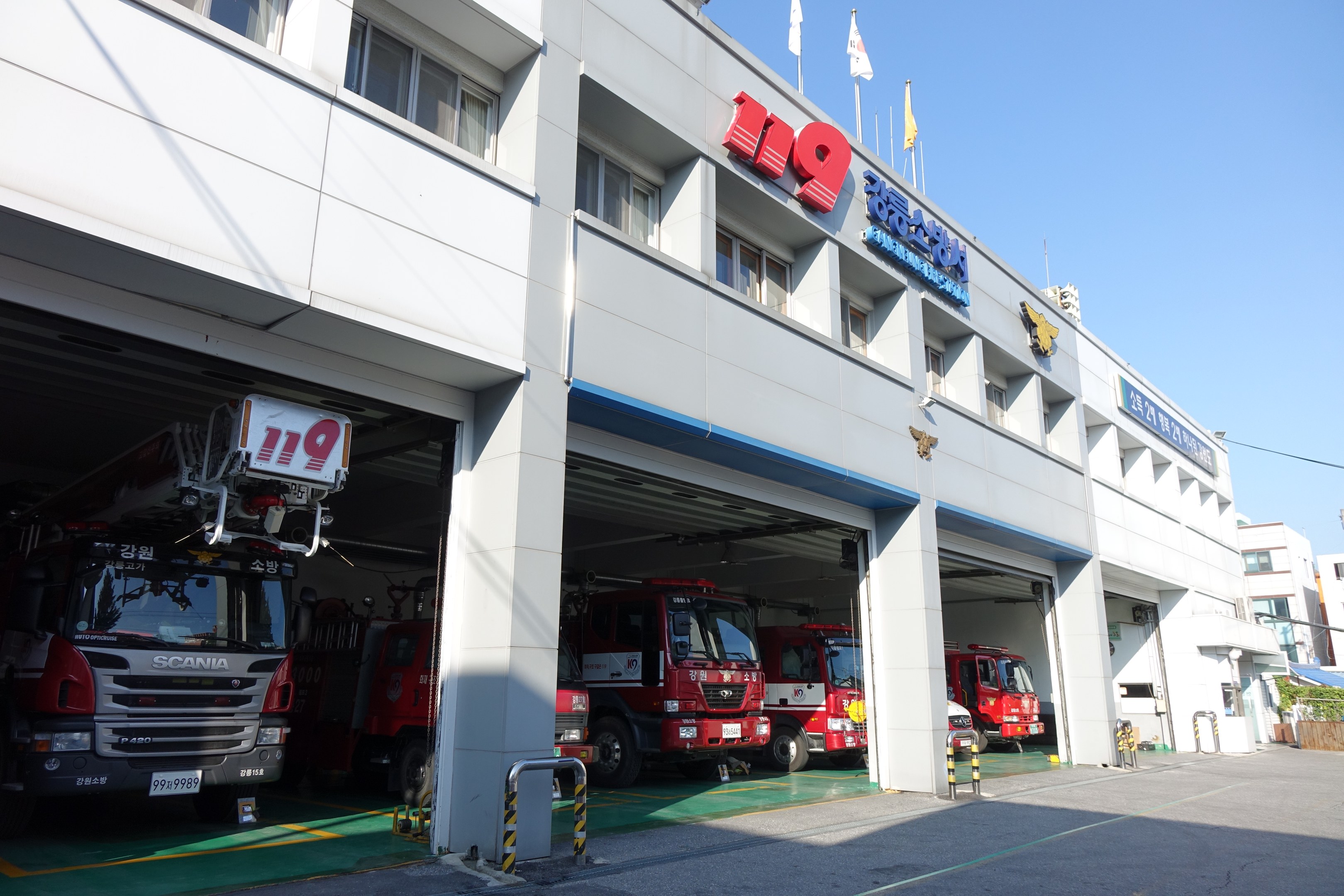 Feuerwache mit mehreren Feuerwehrautos vor dem Gebäude mit Fenstern, Säulen und Namensschildern, mit Flaggen im Vordergrund und anderen Gebäuden vor einem klaren blauen Himmel.