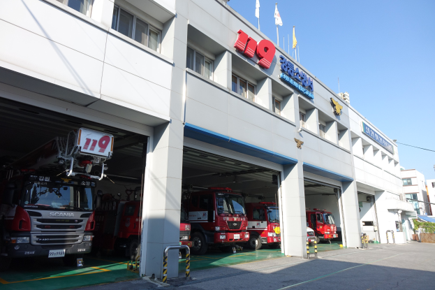 Feuerwache mit mehreren Feuerwehrautos vor dem Gebäude mit Fenstern, Säulen und Namensschildern, mit Flaggen im Vordergrund und anderen Gebäuden vor einem klaren blauen Himmel.