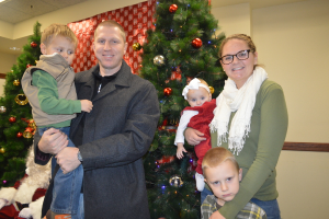 Ein Mann, eine Frau und Kinder stehen vorne mit Weihnachtsbäumen, Santa und einem Plakat im Hintergrund.