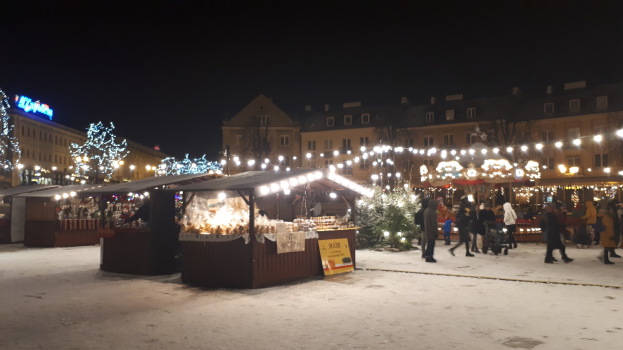 Ein verschneiter Weihnachtsmarkt bei Nacht mit Menschen, Buden mit Lichtern, Pflanzen, Bäumen, Gebäuden und Schildern unter einem bewölkten Himmel.
