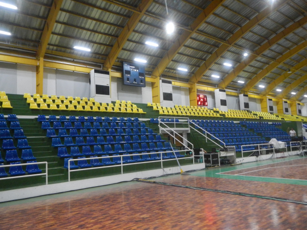 Großer Hallensportplatz mit Holzboden, blauen und gelben Sitzplätzen, Geländern, Treppen mit Geländern, Wandbildschirmen, Deckenleuchten und einigen anwesenden Personen.