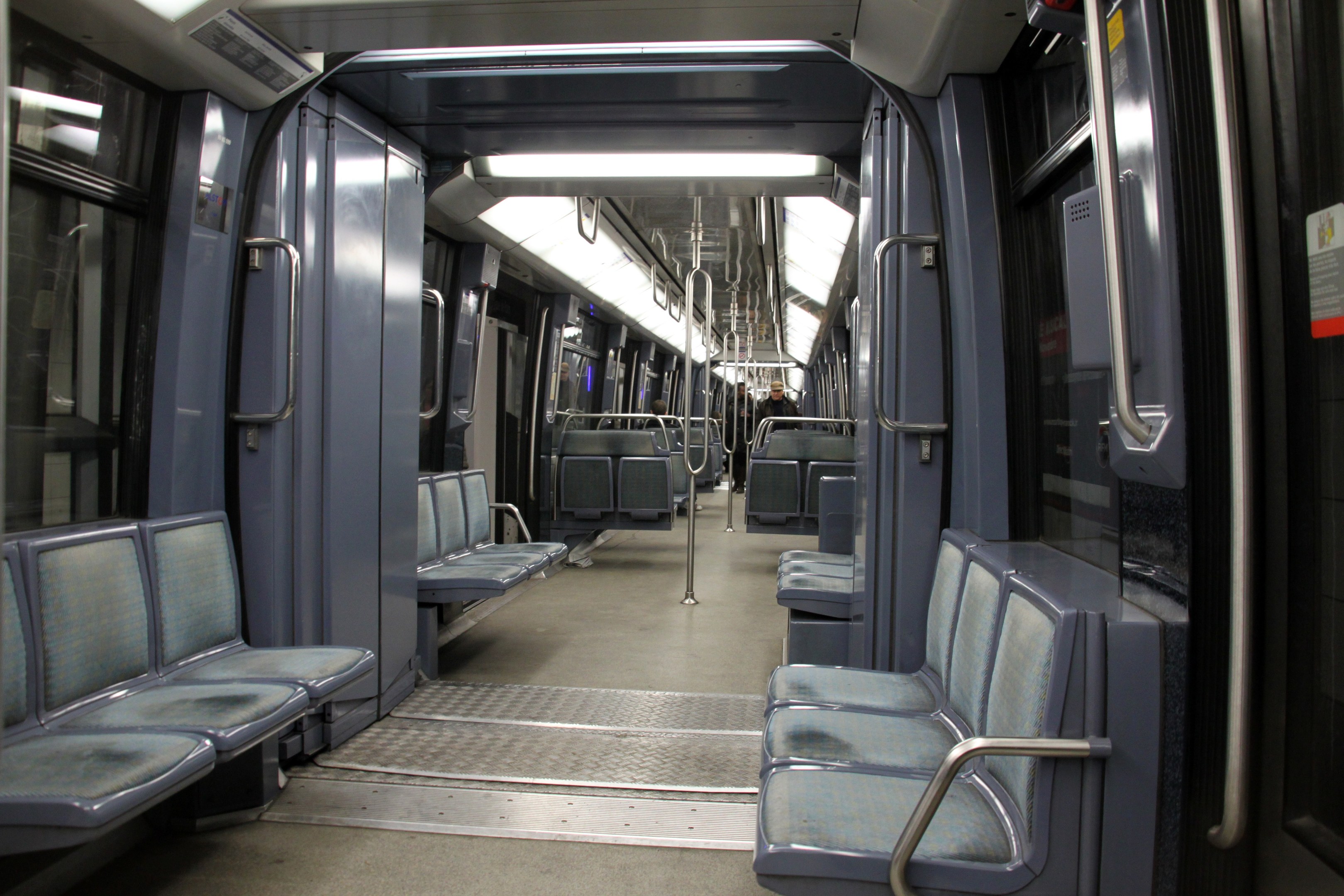 Innenseite eines U-Bahn-Wagens mit blauen Sitzen, Türen, Griffen, Stäben, Lichtern und einigen Menschen in einer belebten Stadtlandschaft.