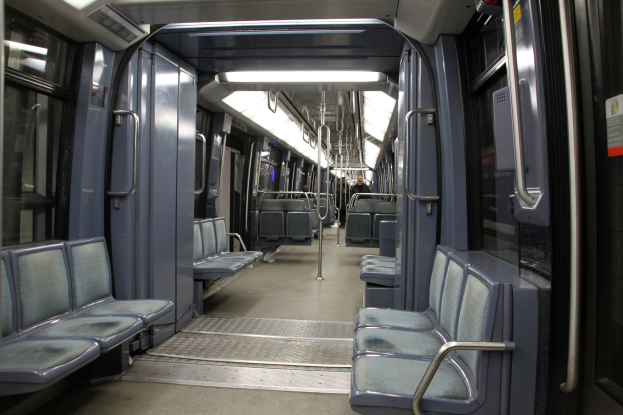 Innenseite eines U-Bahn-Wagens mit blauen Sitzen, Türen, Griffen, Stäben, Lichtern und einigen Menschen in einer belebten Stadtlandschaft.