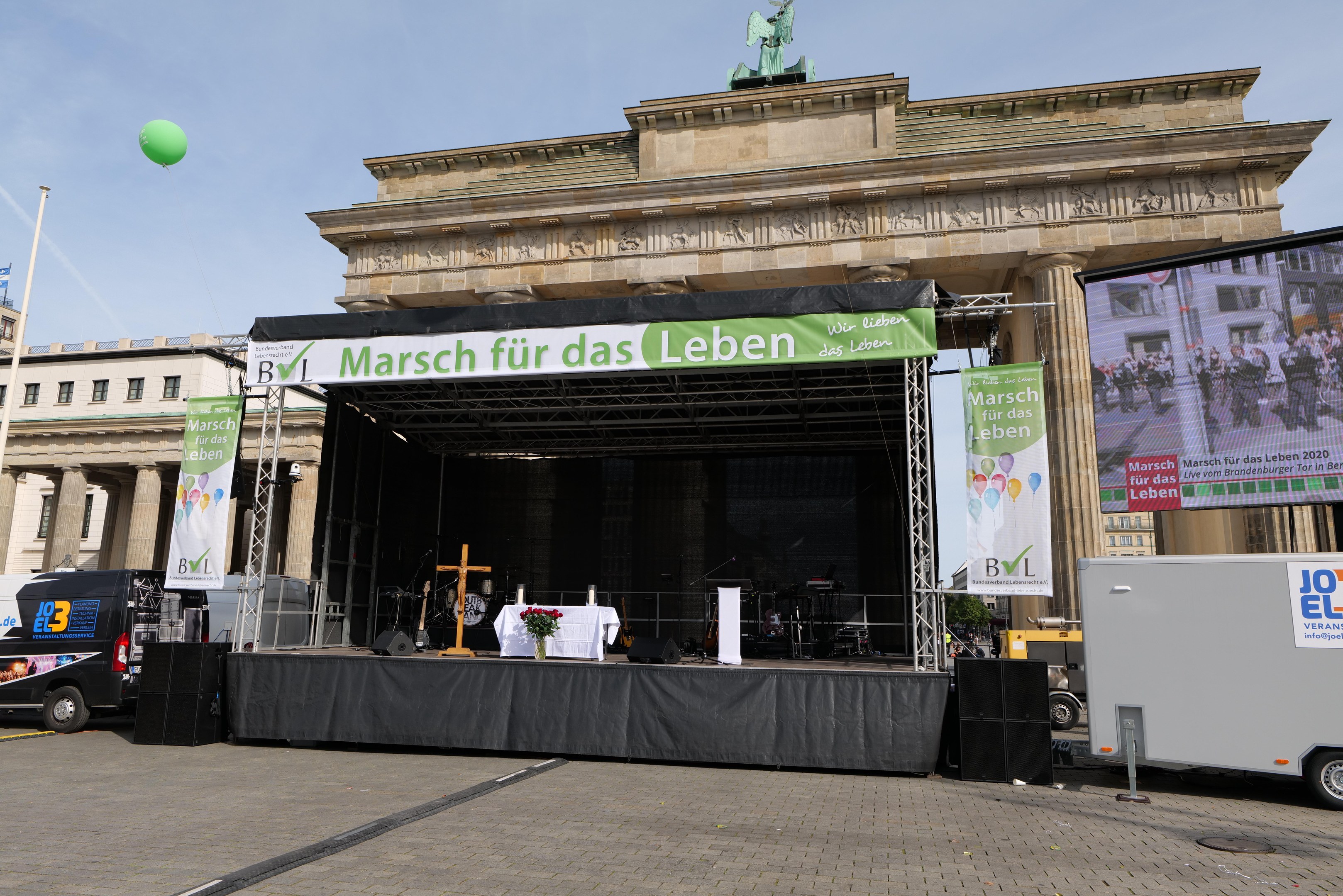 Bühne vor dem Brandenburger Tor mit einem Tisch, Bannern, Lautsprechern, Fahrzeugen, Gebäuden, einer Statue, einer Flagge und bewölktem Himmel.