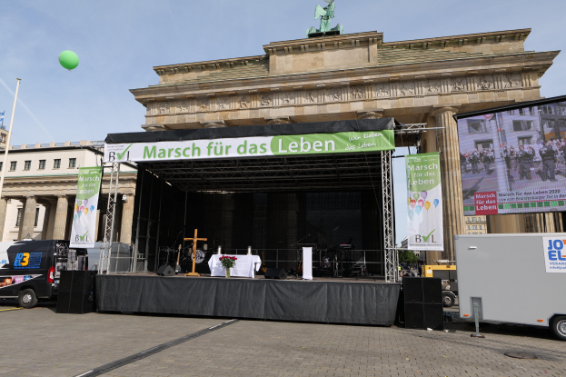 Bühne vor dem Brandenburger Tor mit einem Tisch, Bannern, Lautsprechern, Fahrzeugen, Gebäuden, einer Statue, einer Flagge und bewölktem Himmel.