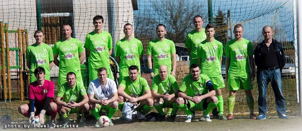 Mannergruppe in grünen T-Shirts auf einem Fußballfeld mit einem Ball davor, einige im Squat, in der Nähe eines Torpfostens mit Bäumen und Gebäuden im Hintergrund.