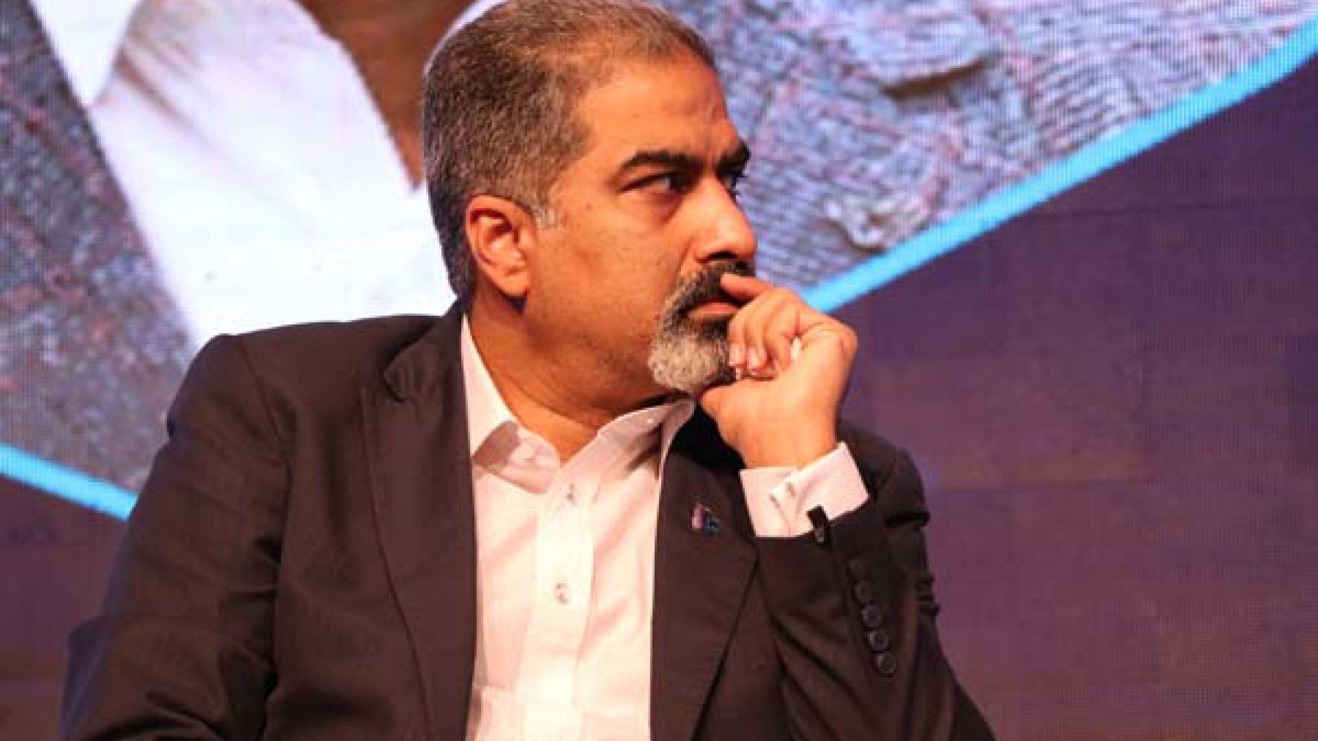 Ein Mann in Anzug und Krawatte sitzt auf einer Bühne und schaut nachdenklich auf einen Bildschirm.