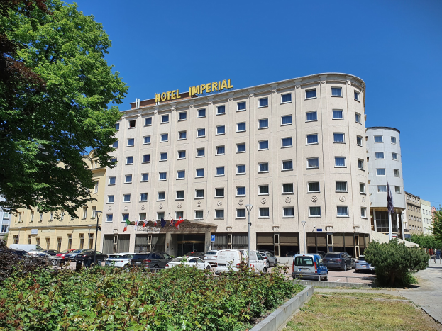 Großes beiges Gebäude mit der Aufschrift "Imperial Hotel" und Fenstern, umgeben von Grün, parkenden Autos, flaggengeschmückten Pfählen und einem bewölkten Himmel.