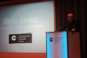 Eine Person steht auf einem Podium und hält eine Rede, vor ihr befinden sich zwei Mikrofone und ein großer Bildschirm zur Seite.