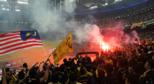Eine große Menge Menschen in einem Stadion mit brennenden Fackeln in der Luft, einige halten Fahnen und eine Person hält eine Kamera, mit Rauch im Hintergrund und Schildern mit der Aufschrift "Malaysia vs Malaysia Live Streaming".