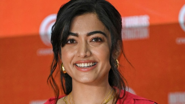 Eine Frau in einem roten Kleid mit einer eleganten Hochsteckfrisur und dezentem Make-up lächelt in die Kamera, trägt schwarze High Heels und hat ein hellgelbes Banner mit der Aufschrift 'Die besten Schönheits Looks von Sonam Kapoor beim Cannes Film Festival' im Hintergrund.