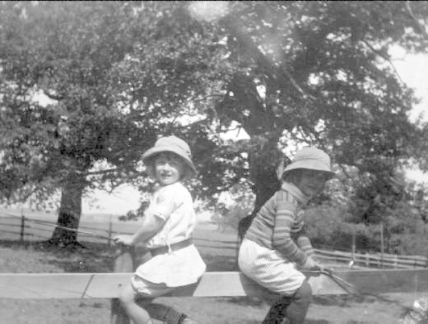 Zwei junge Jungs in Mützen halten Baseballschläger in einem grasbewachsenen Park mit Bäumen und einem Zaun im Hintergrund, in Schwarz-Weiß.
