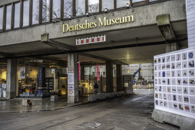 Außenansicht des Deutschen Museums in Berlin, das einen großen Glaspavillon mit Säulen, ein Namensschild und eine Informationsanzeigetafel auf der rechten Seite sowie Bäume und andere Gebäude im Hintergrund zeigt.