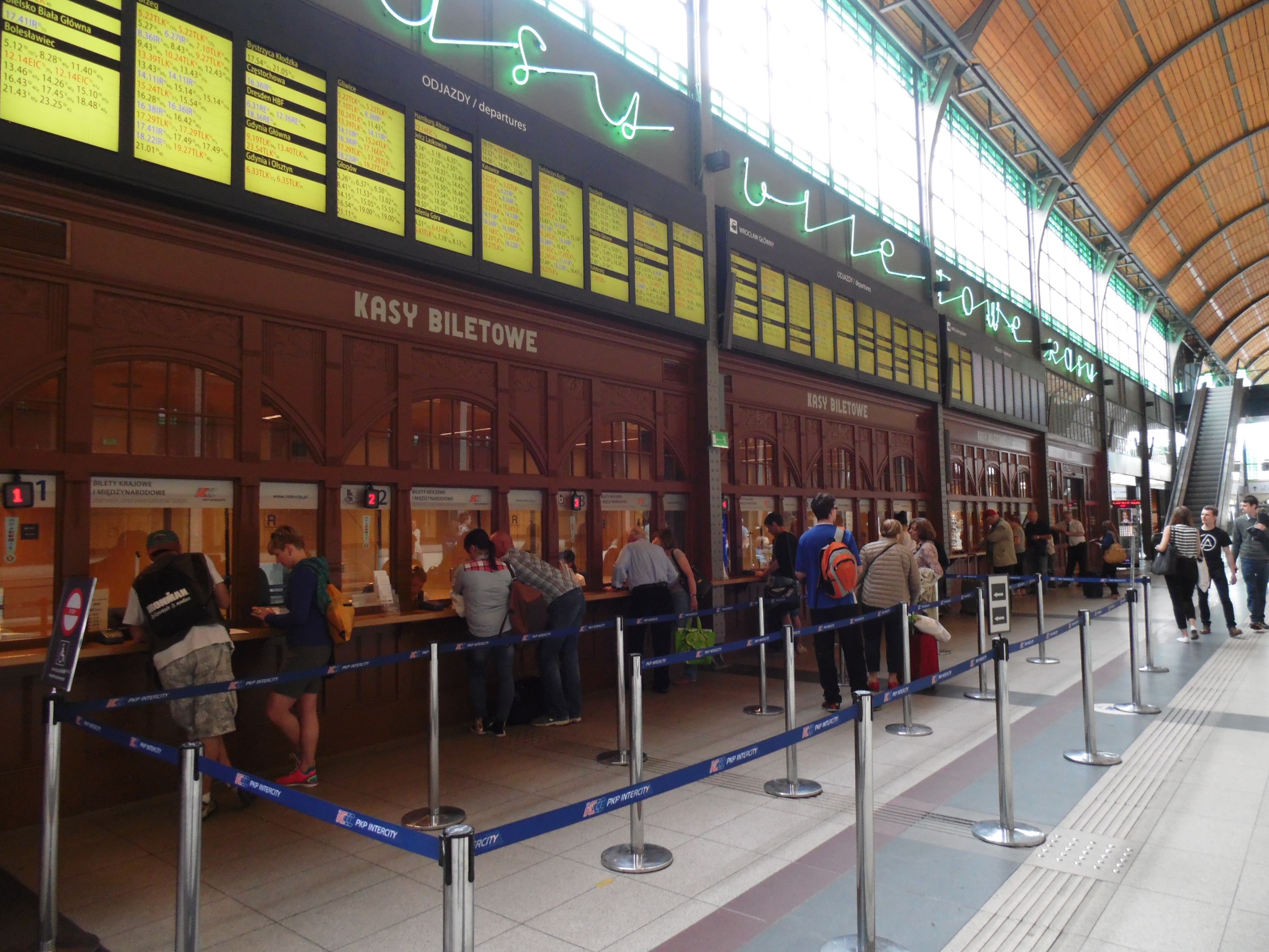 Gruppe von Menschen an einem Bahnhof-Schalter mit Gepäck, Stangen mit Bändern, Infotafeln, Deckenbeleuchtung und Treppen mit Geländern im Hintergrund.