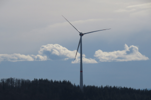Windkraftanlage in einem Wald mit bewölktem Himmel und Bergen im Hintergrund.