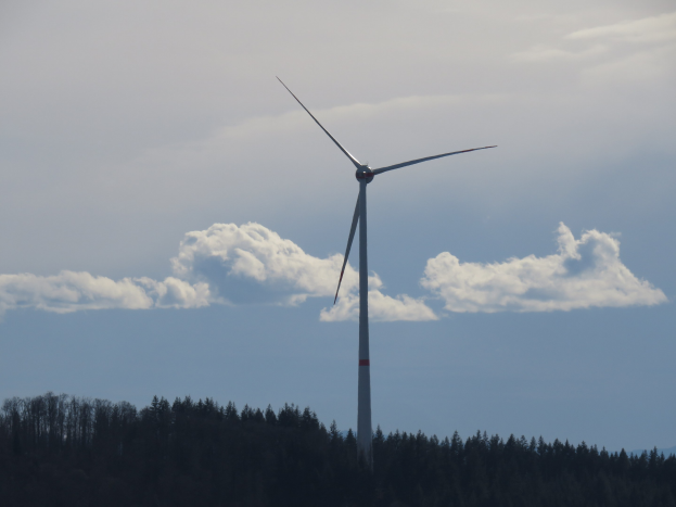 Windkraftanlage in einem Wald mit bewölktem Himmel und Bergen im Hintergrund.