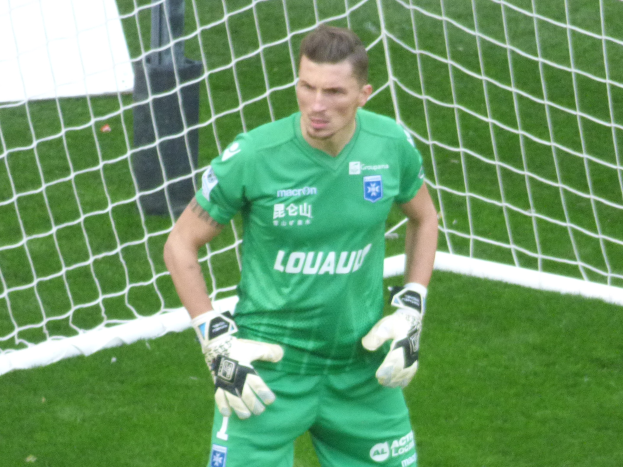Ein Torwart in grüner Uniform steht vor einem Fußballtor, trägt Handschuhe auf einem grünen Rasen.