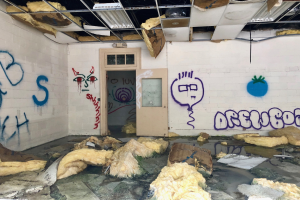 Ein Raum mit Graffiti an den Wänden, eine Tür im Hintergrund, Deckenstangen und Lampen sowie verschiedene Gegenstände, die auf dem Boden verstreut sind.