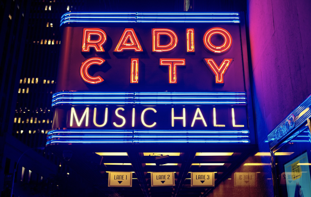 Außenansicht des Radio City Music Hall in New York City bei Nacht, hell erleuchtet und von hohen Gebäuden flankiert, mit einem "Radio City"-Schild und zusätzlichen Tafeln auf der rechten Seite.