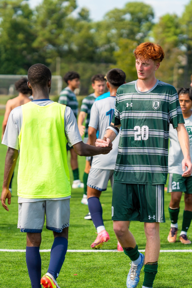 Junge Männer auf einem Fußballfeld mit Turnschuhen, umgeben von Bäumen und einem klaren blauen Himmel.