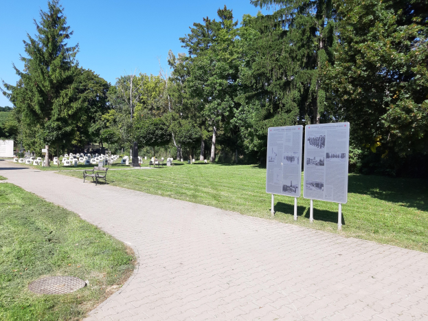 Ein Park mit einem zentralen Gras- und Baumgesäumten Weg, der von einer Bank im Vordergrund, Informationsschildern und Gräbern im Hintergrund unter einem klaren blauen Himmel umgeben ist.