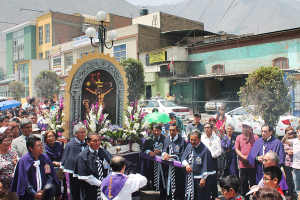 Menschen auf einer Stra├če vor der Stadt mit einem Kruzifix und Blumenstr├Ąu├čen im Vordergrund, fahrende Autos im Mittelgrund und Geb├Ąude, B├Ąume und Stra├čenlaternen im Hintergrund.