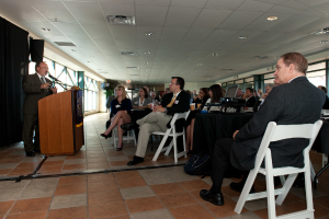 Ein Mann steht an einem Podium und spricht in ein Mikrofon, während vor ihm eine Gruppe von Männern und Frauen sitzt und im Hintergrund Fenster zu sehen sind.