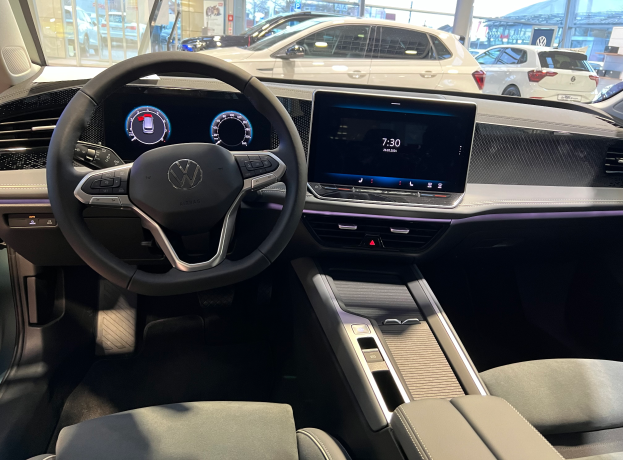 Das Innere eines Volkswagen e-Golf mit Lenkrad, Armaturenbrett, Bildschirmen, Knöpfen, Sitzen und Glastüren, mit Blick auf andere Autos, Gebäude, Bäume, Pfosten und den Himmel.