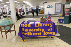Eine Ausstellung mit einem Tisch, auf dem verschiedene Landesflaggen, ein Karton, Papiere und ein Banner sowie links daneben eine Weltkugel, rechts daneben eine Anzeigetafel mit einigen Menschen in der Nähe und mehrere Tische und Stühle im Hintergrund zu sehen sind.