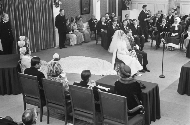 Schwarze und weiße Fotografie einer Hochzeitszeremonie mit sitzenden und stehenden Menschen, einem Mann mit Mikrofon, einer Frau mit einem Blumenstrauß, einem Podium mit Mikrofon, Blumengestecken, einem Vorhang, einem gerahmten Foto an der Wand und einer Tür im Hintergrund.
