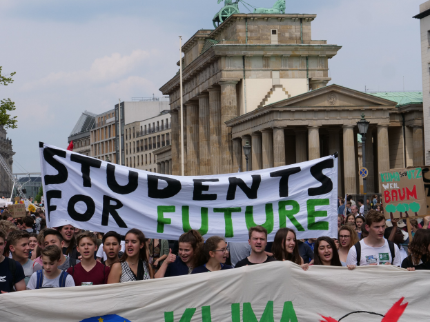 Eine Gruppe von Studenten marschiert in Berlin, eine bunt bemalte Fahne schwingend, auf der 'Students for Future' steht, mit Gebäuden, Bäumen und Himmel im Hintergrund.