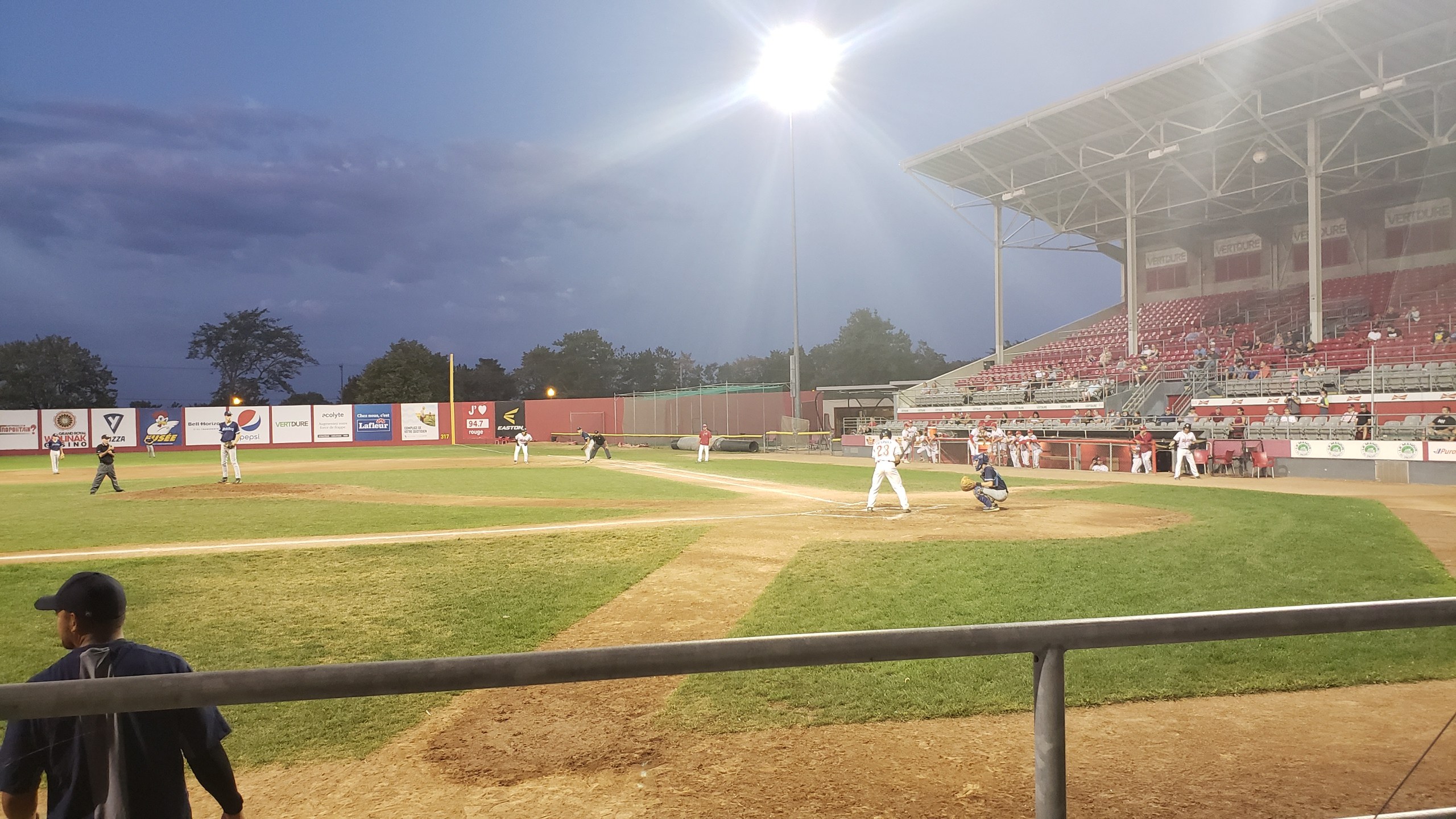 Baseballspiel im Gange mit Zuschauern auf den Tribünen, Gelúnders im Vordergrund, Bürgen, Masten, Lichter, Werbetafeln und wolkenlosem Himmel im Hintergrund.