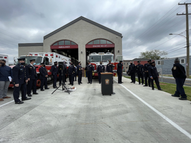 Gruppe von Menschen in Mützen neben einem Feuerwehrauto auf einem Podium während einer Feuerwache-Einweihungszeremonie, mit Gebäuden, Bäumen, Strommasten, Gras und einem bewölkten Himmel im Hintergrund.