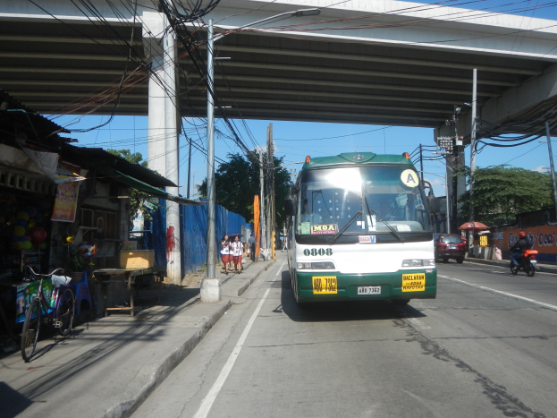 Ein grüner und weißer Bus fährt eine Straße entlang, die von Strommasten und -leitungen gesäumt ist, vorbei an Geschäften, Ständen, parkenden Fahrrädern und Fußgängern, mit einer Brücke und Bäumen im Hintergrund bei einem klaren blauen Himmel.