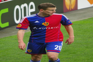 Ein Mann in einem roten und blauen Fußballtrikot, identifiziert als Flügelspieler, läuft auf dem Feld mit ausgestreckten Armen und entschlossener Miene, während einige Menschen auf den Tribünen hinter ihm sitzen.