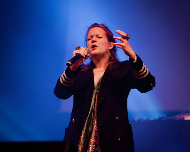Eine Frau in einem schwarzen Kleid mit einem Dutt singt leidenschaftlich auf einer Bühne unter blauer Beleuchtung, hält ein Mikrofon und hat die Augen geschlossen.