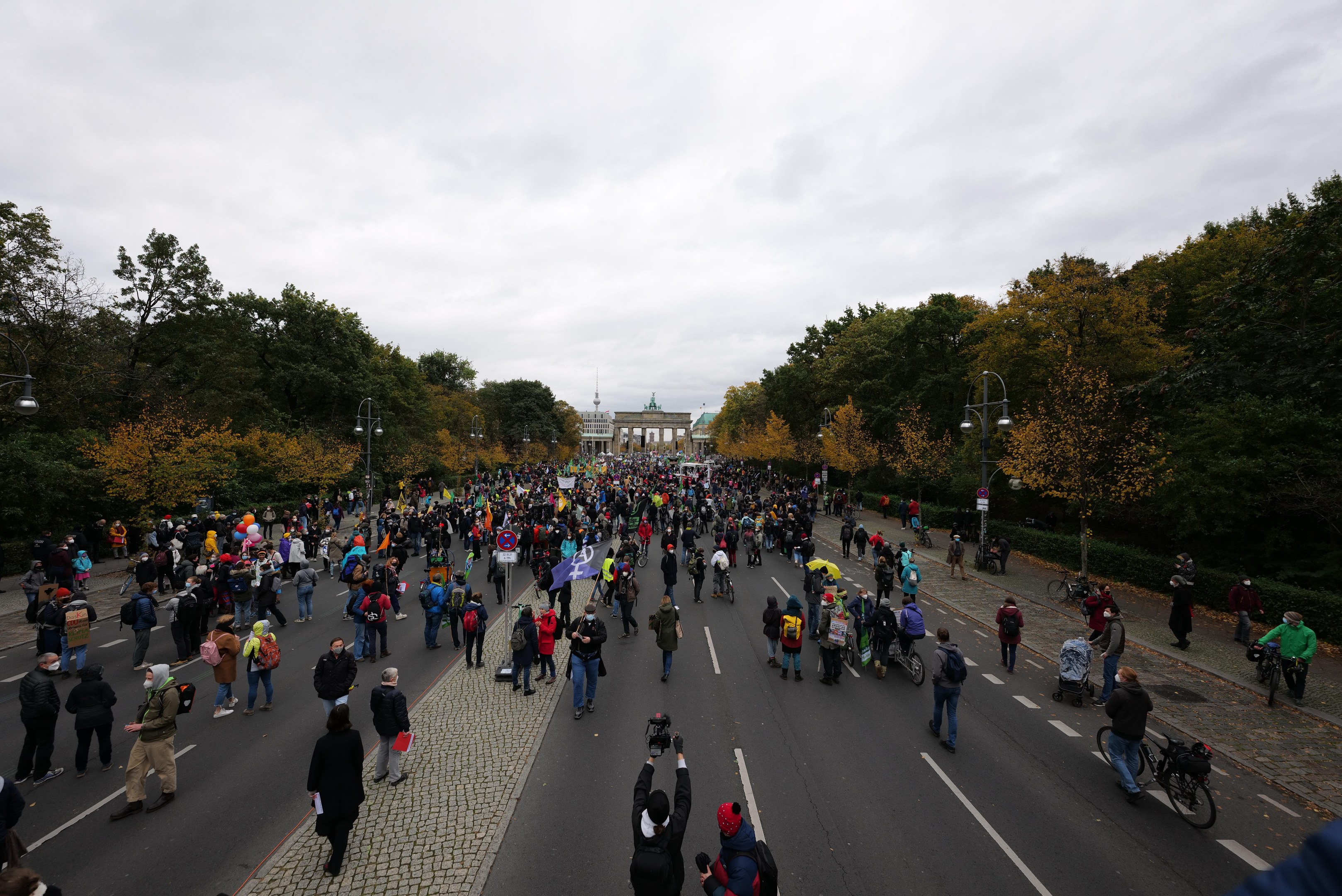 Eine große Gruppe von Menschen marschiert auf einer von Bäumen gesäumten Straße während einer Protestdemo in Berlin, wobei einige Kameras halten und ein Gebäude im Hintergrund unter einem klaren Himmel zu sehen ist.