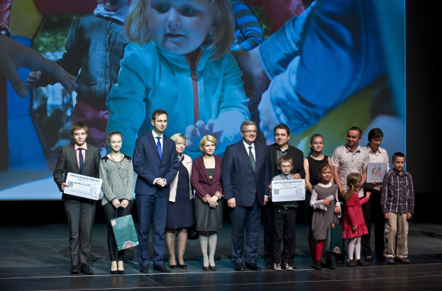 Gruppe von Menschen auf einer Bühne mit Taschen und Urkunden, mit einem Bildschirm im Hintergrund, der ein Kind zeigt.